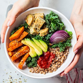Woman hands eating vegan salad of baked vegetables, avocado, tofu and buckwheat buddha bowl, top view. Plant based food concept.